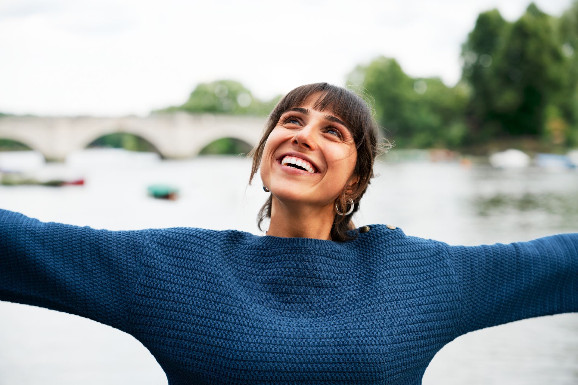 Woman smiles with arms outstretched near a river and bridge; she wears a blue sweater.