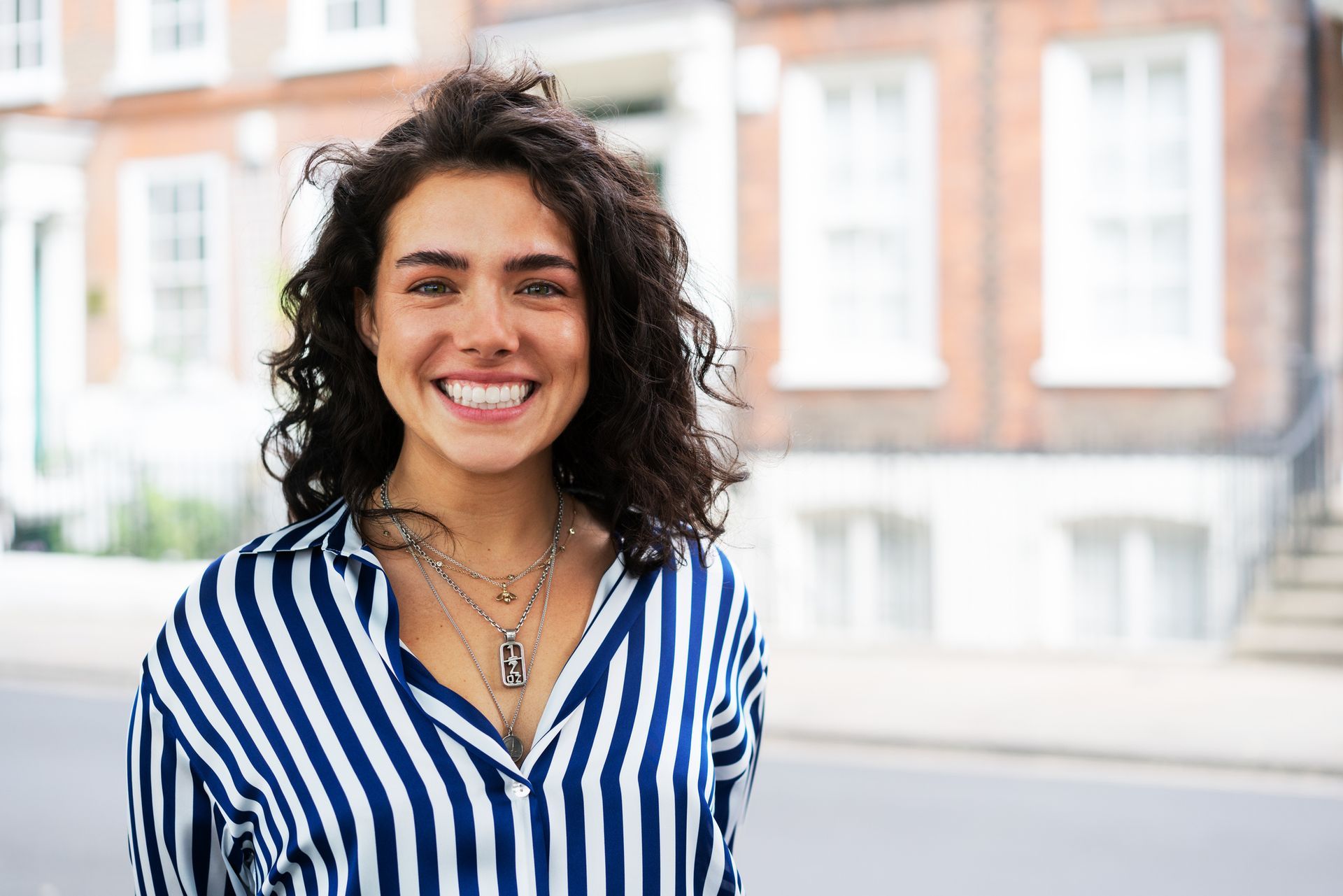 Woman with curly hair smiles, wearing a blue and white striped shirt; street and brick building background.