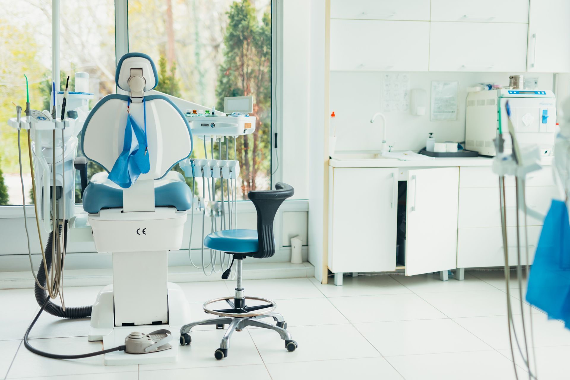 Dentist's office with blue chair, equipment, and patient chair. Bright, white room.