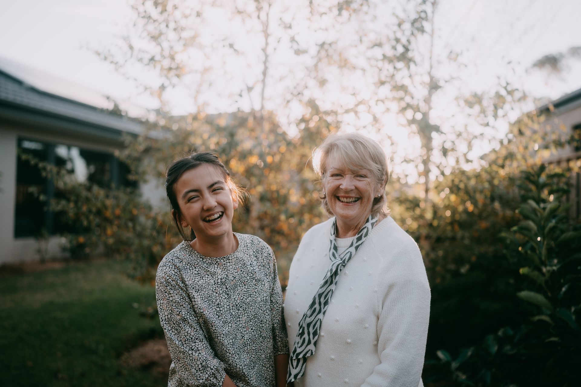 Two smiling women pose in a sunny garden. One wears a patterned top; the other, a white sweater and scarf.