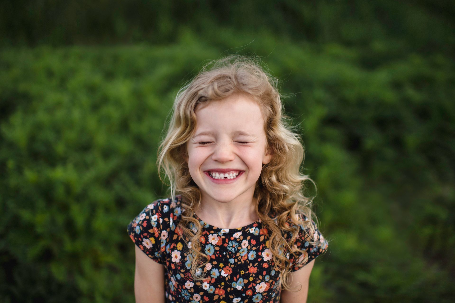 Girl with curly blonde hair smiles widely, eyes closed, in front of a green, leafy background.