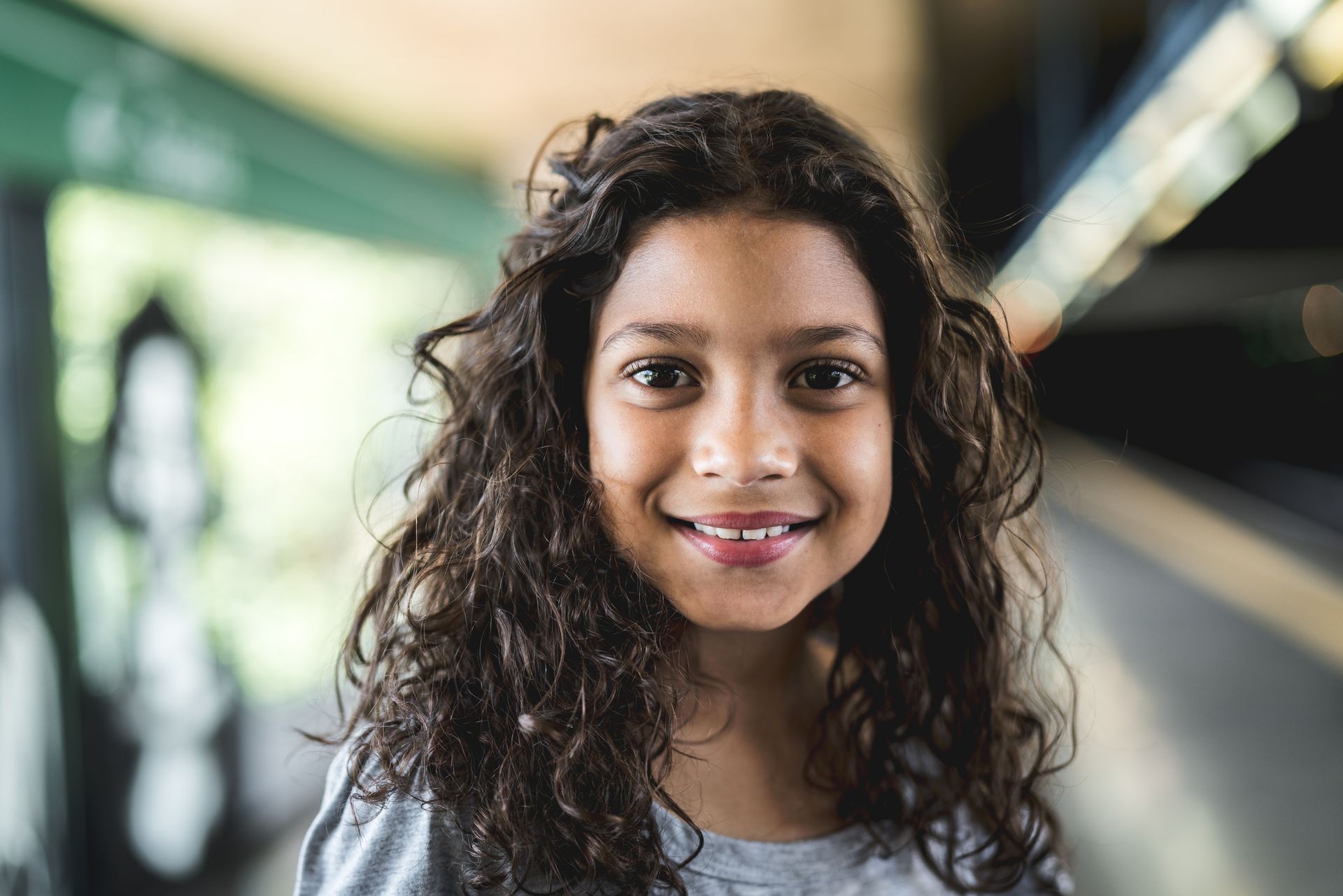 Smiling girl with curly brown hair; blurred background of a public transit station.