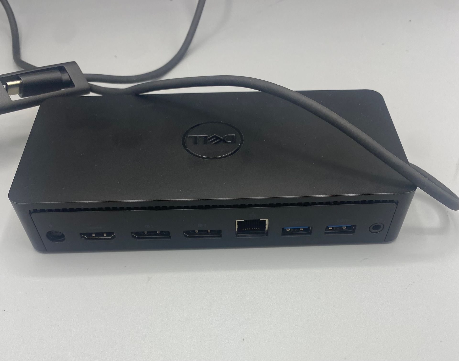Black Dell docking station with attached USB cable, lying on a white surface.