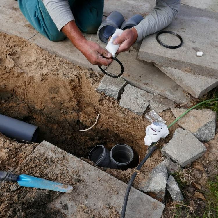 Worker installing black cables and PVC pipes in a trench beside broken concrete slabs.