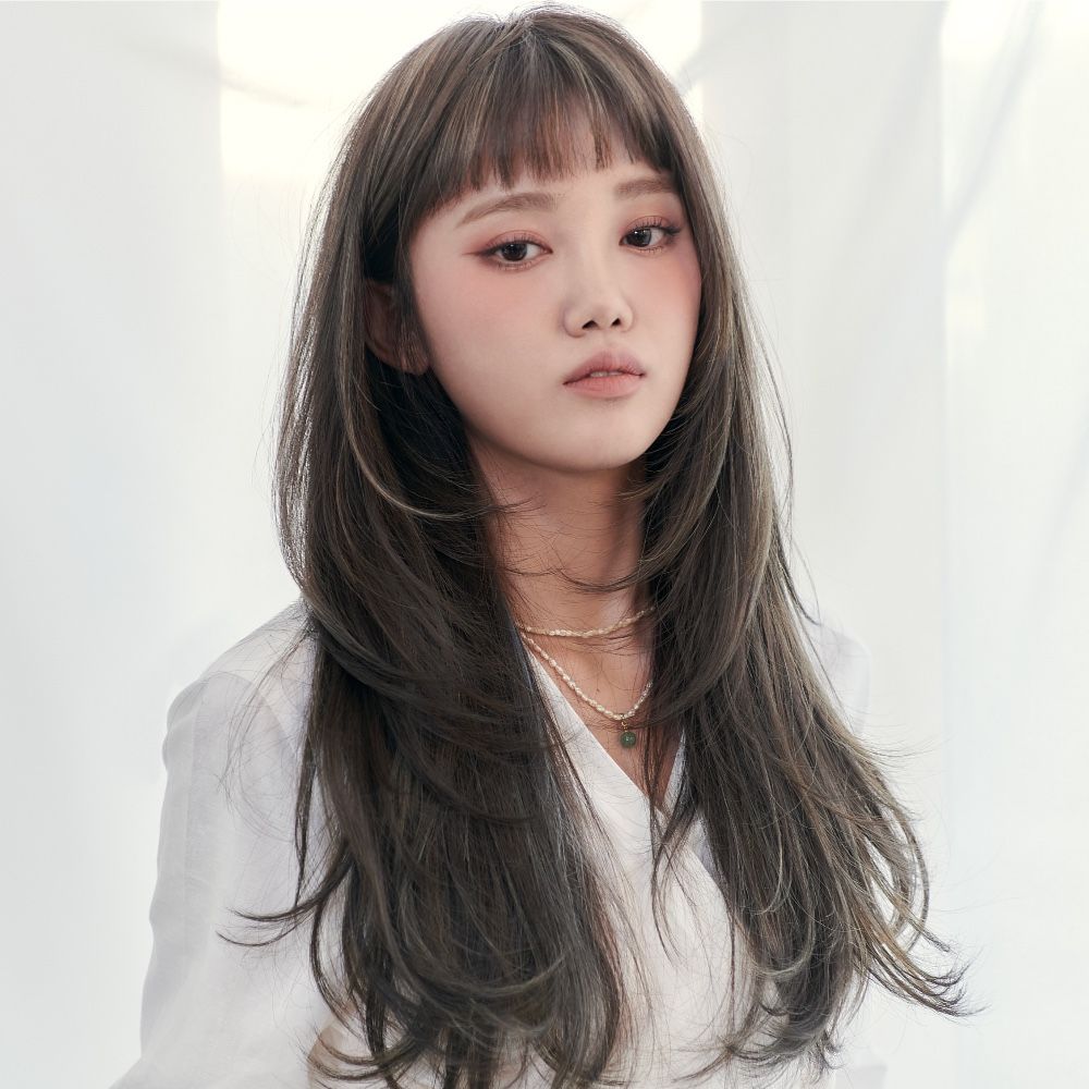 A woman with long hair and bangs is wearing a white shirt and a necklace.