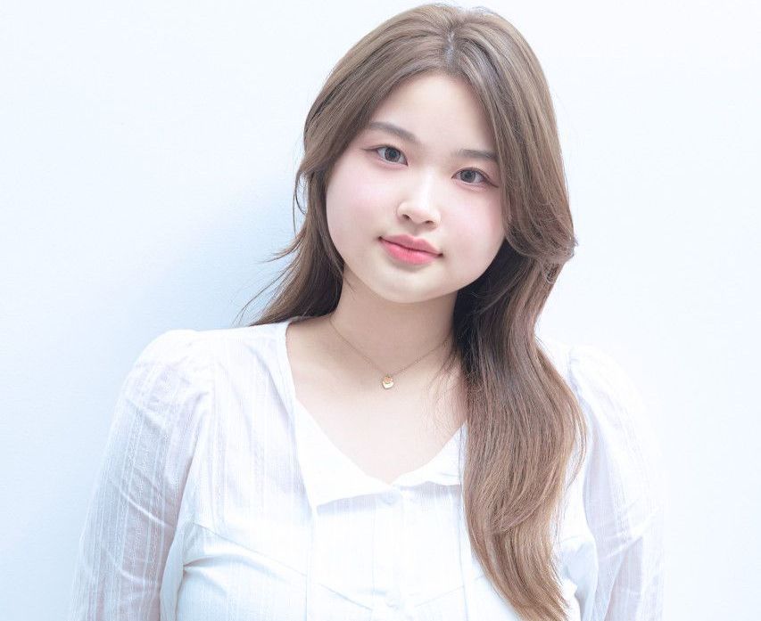 A young woman wearing a white shirt and a necklace is standing in front of a white wall.