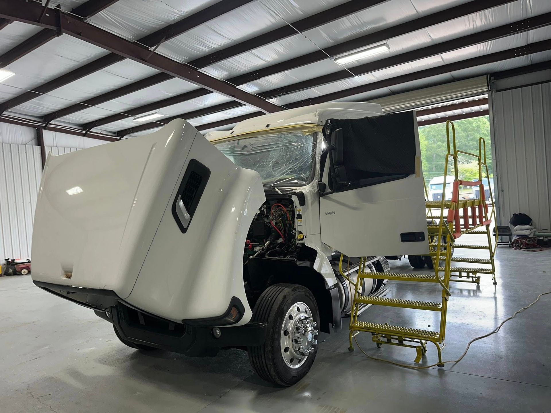 White truck with hood open, parked in a shop. A yellow scaffold is alongside the cab.