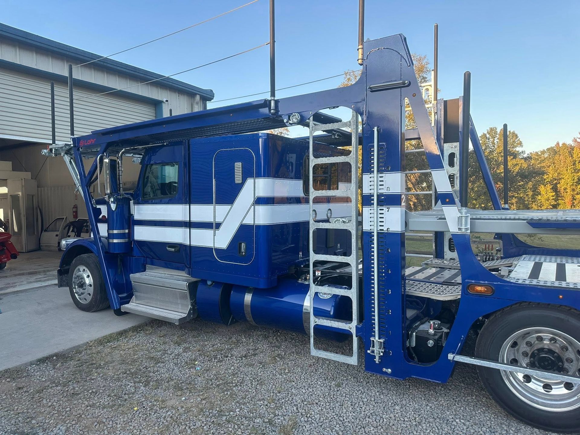 Blue car hauler truck parked by a building, featuring white stripes and a side ladder.