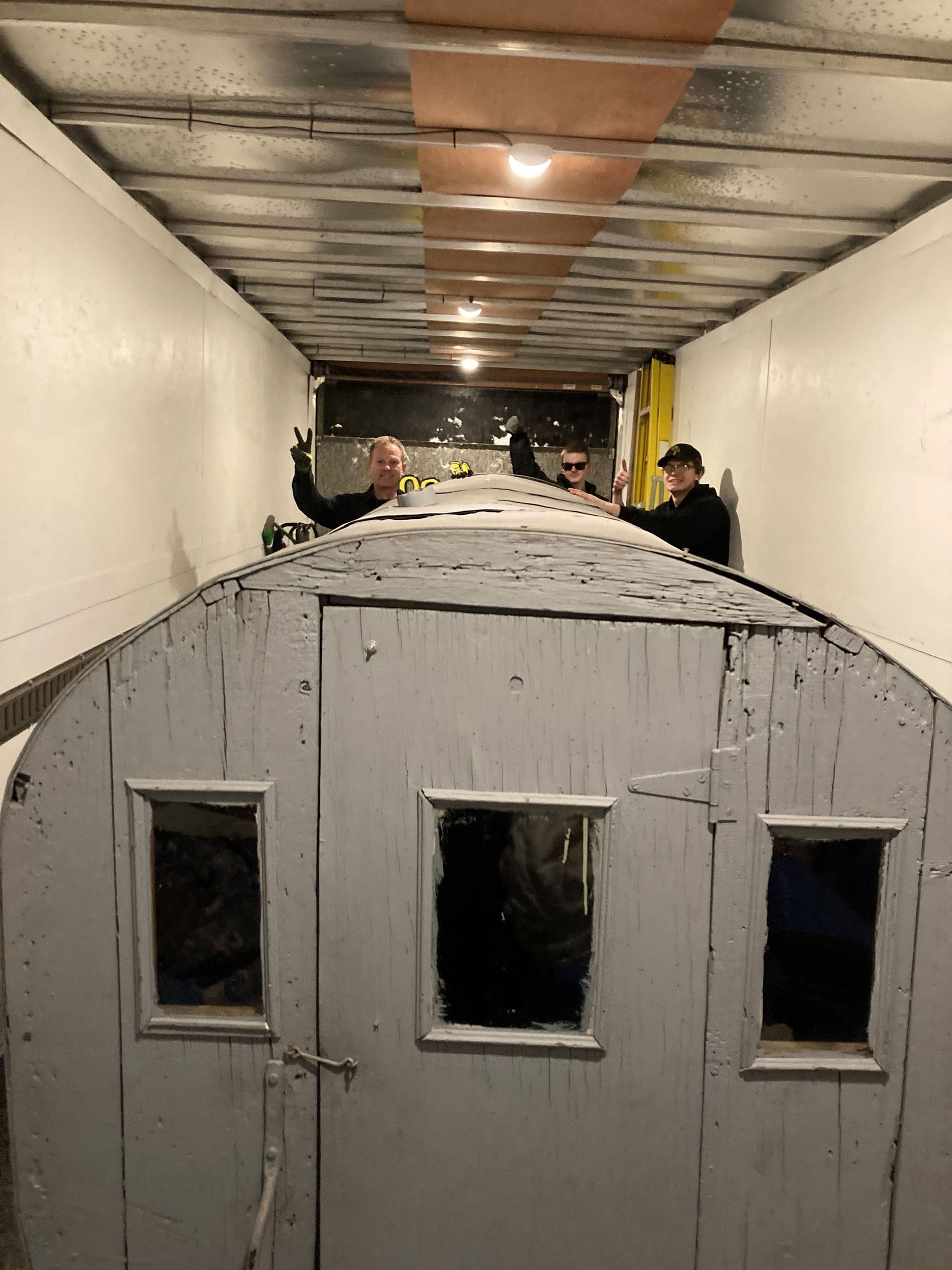 Three people work on a white, weathered, wooden structure inside a trailer.