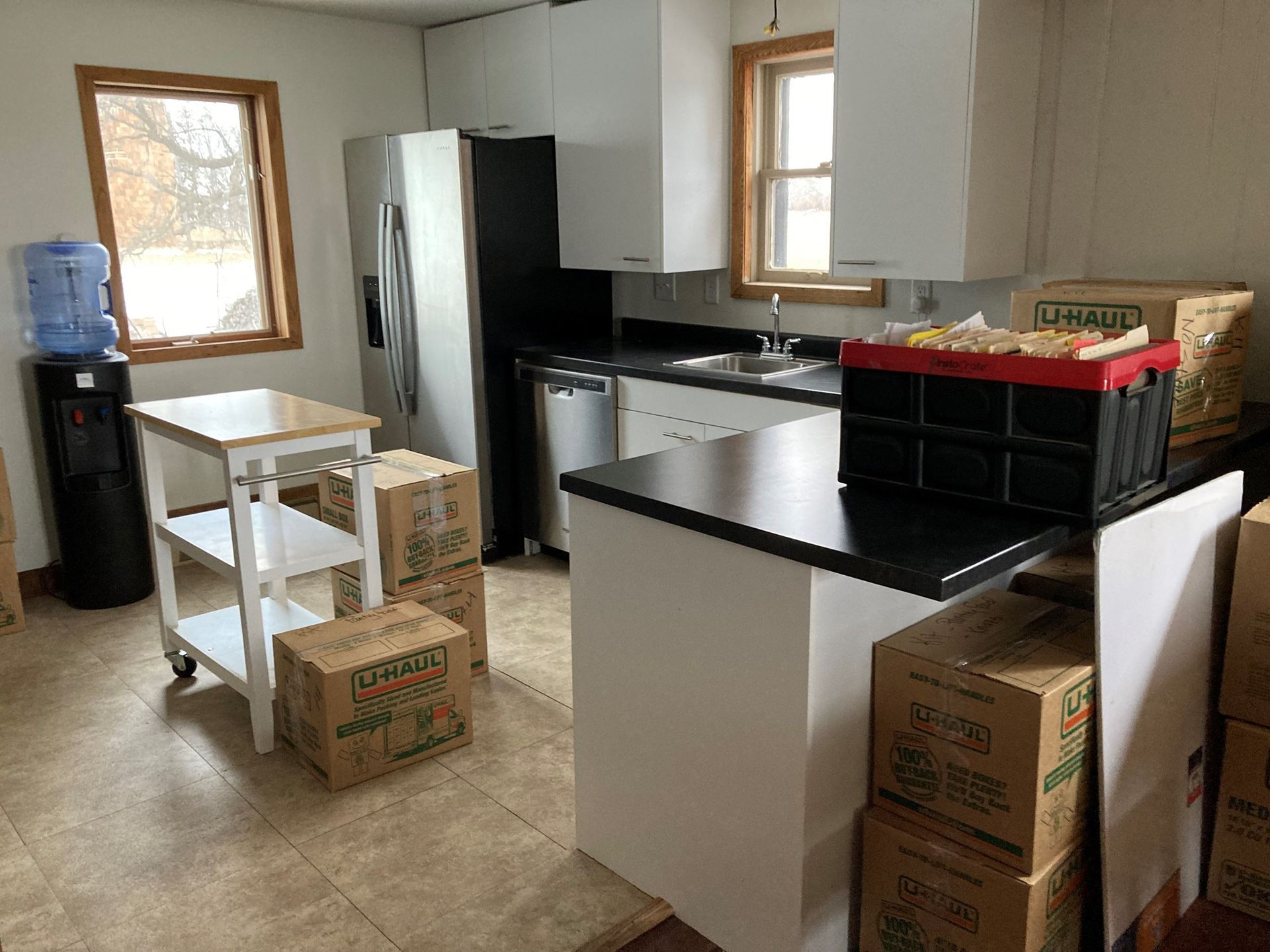 Kitchen with white cabinets, black countertops, refrigerator, island, boxes, water dispenser, and rolling cart.