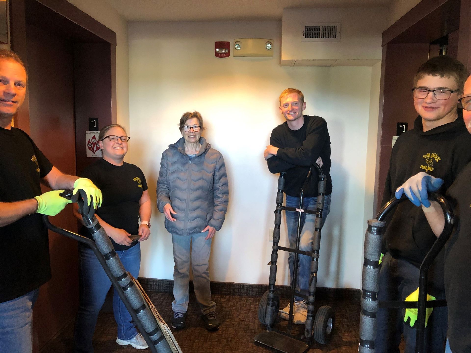 People with hand trucks in an elevator lobby; two elevators, one with a person in the center wearing a gray jacket.