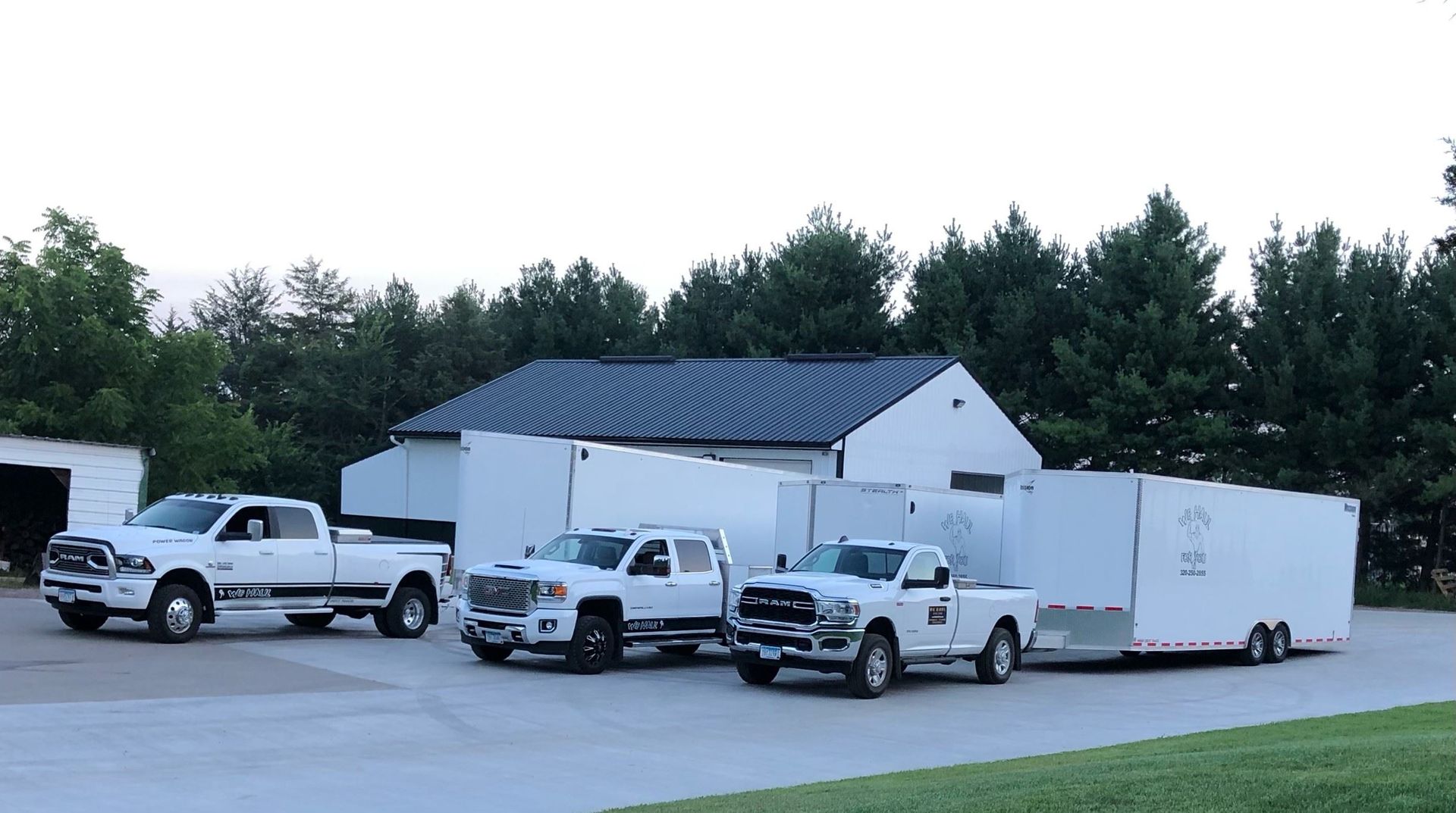 Three white pickup trucks and a trailer parked in front of a white building, set against a backdrop of trees.