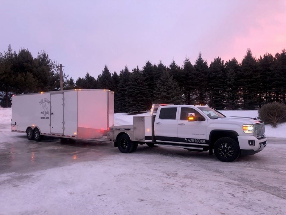 White truck with a trailer parked on a snowy surface in front of a forest at dusk.