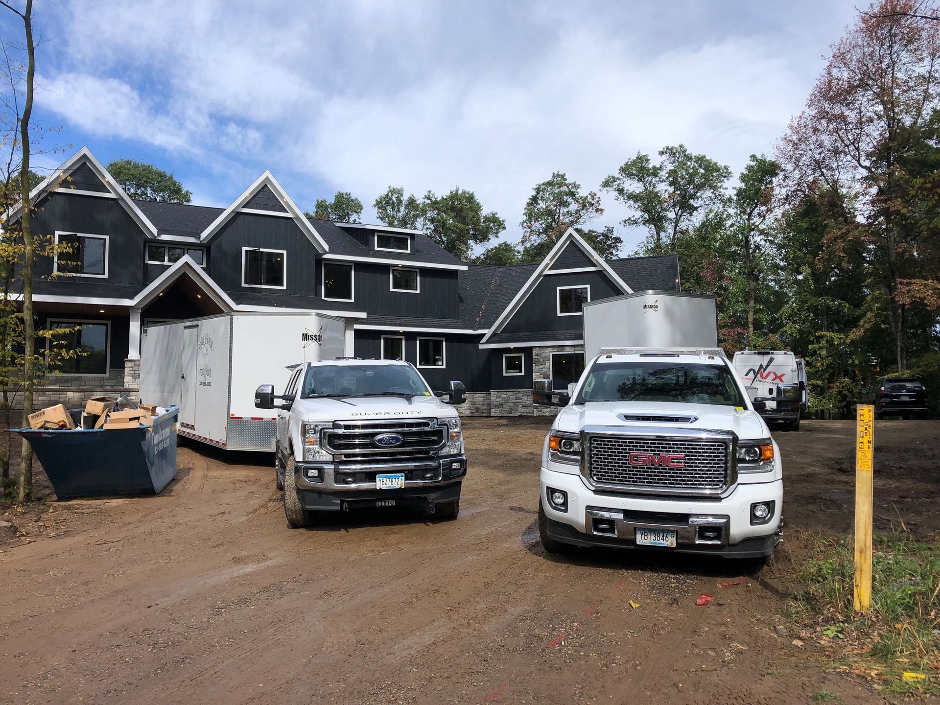 Two trucks parked in front of a dark house under construction, with a trailer and dumpster.