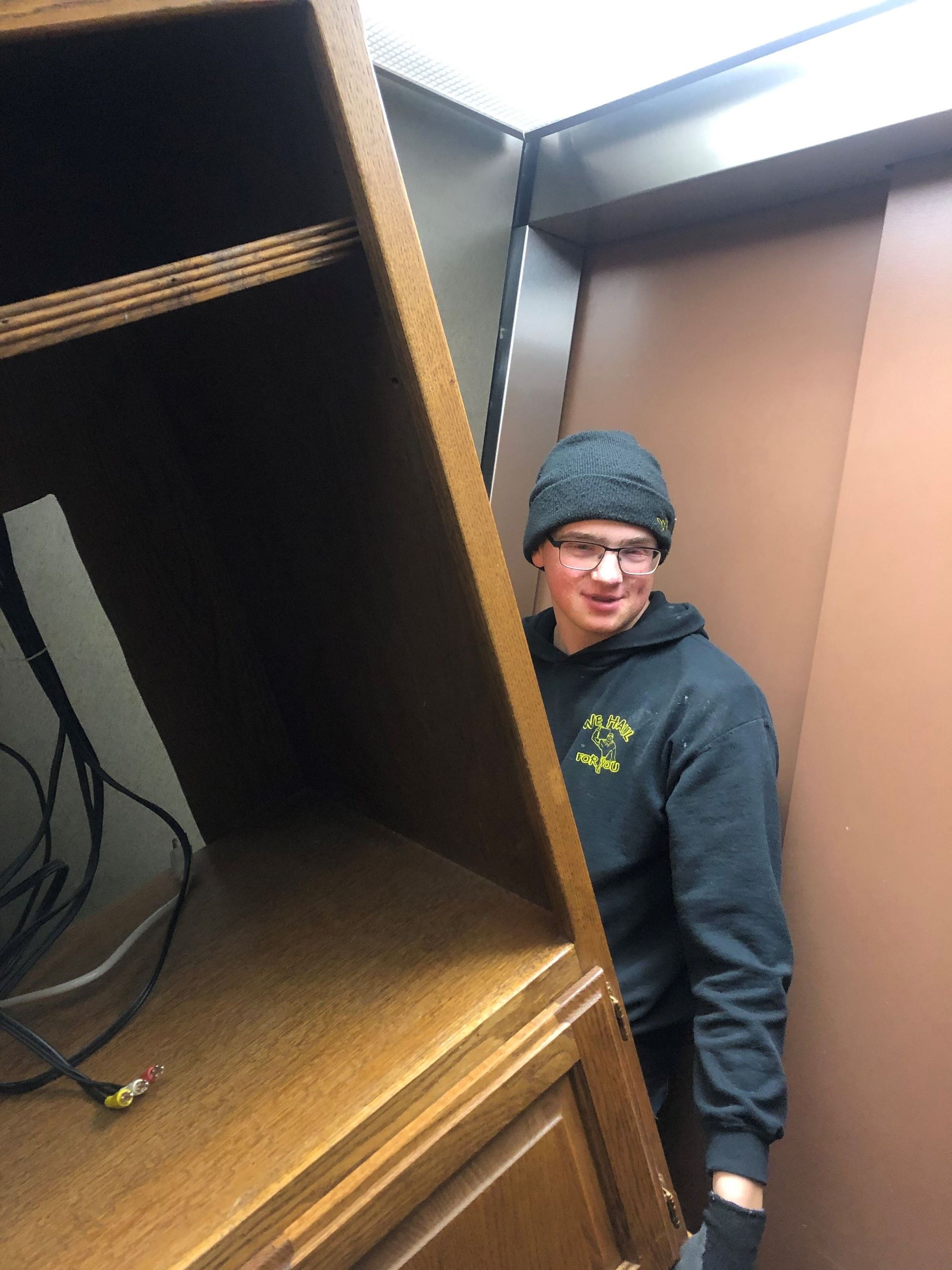 Person carrying a wooden cabinet in an elevator, smiling. They wear a beanie, glasses, and a dark sweatshirt.