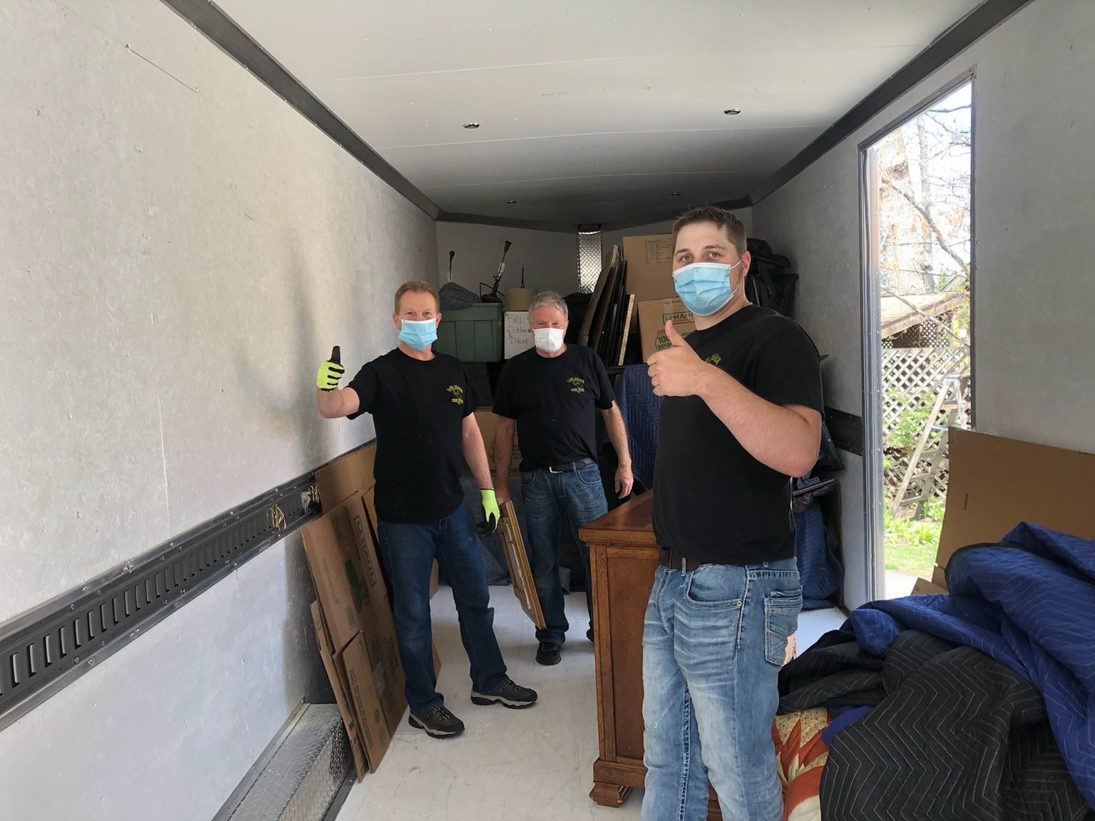 Three movers in masks inside a trailer, loading furniture and boxes. One gives a thumbs-up.