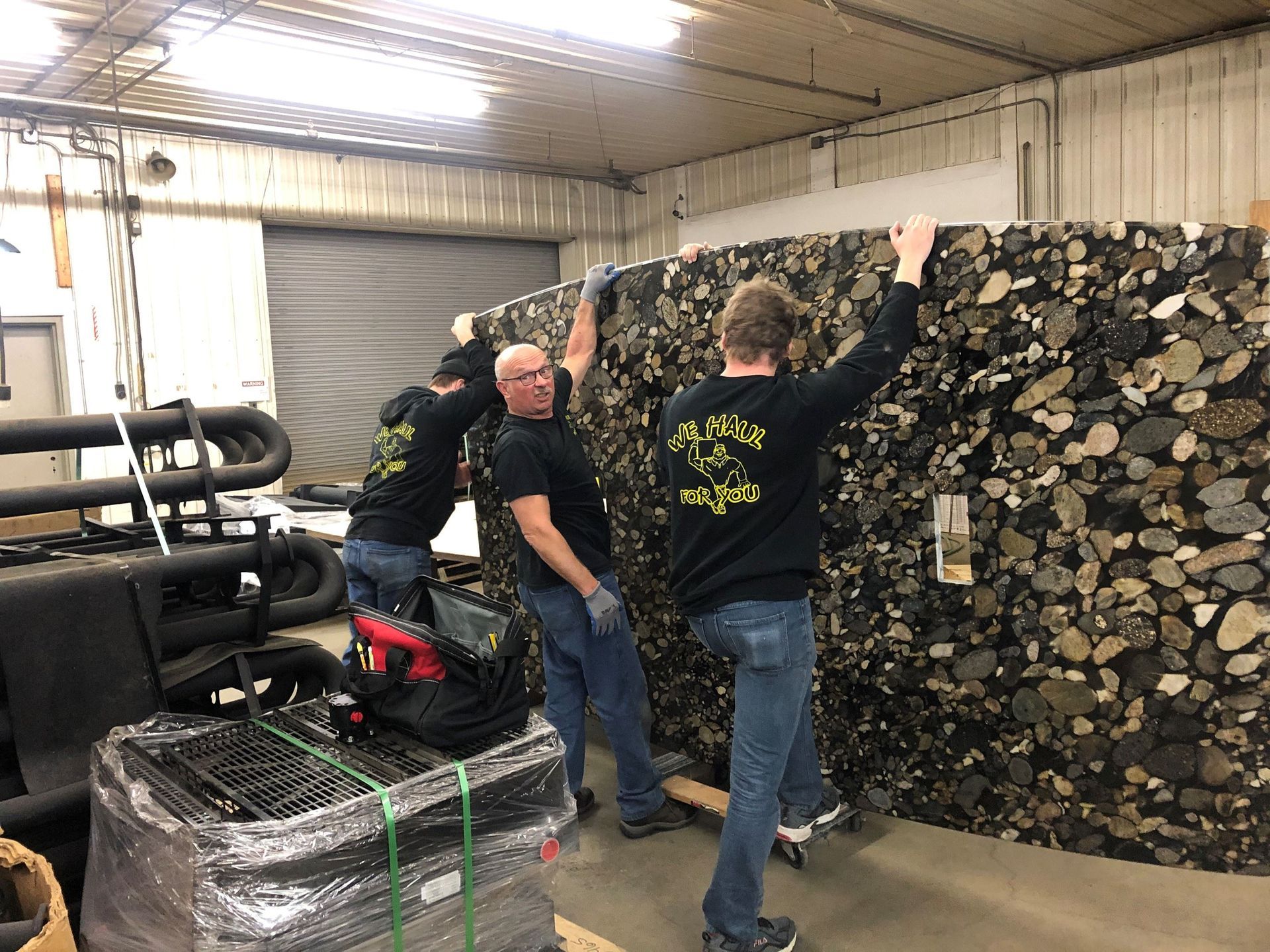 Three people lift a large, dark foam slab indoors. Two are on the left, one on the right, all wearing black shirts, jeans.