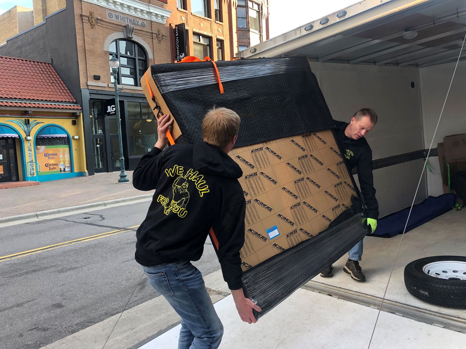 Two people loading large, cardboard-wrapped object into a truck on a city street.