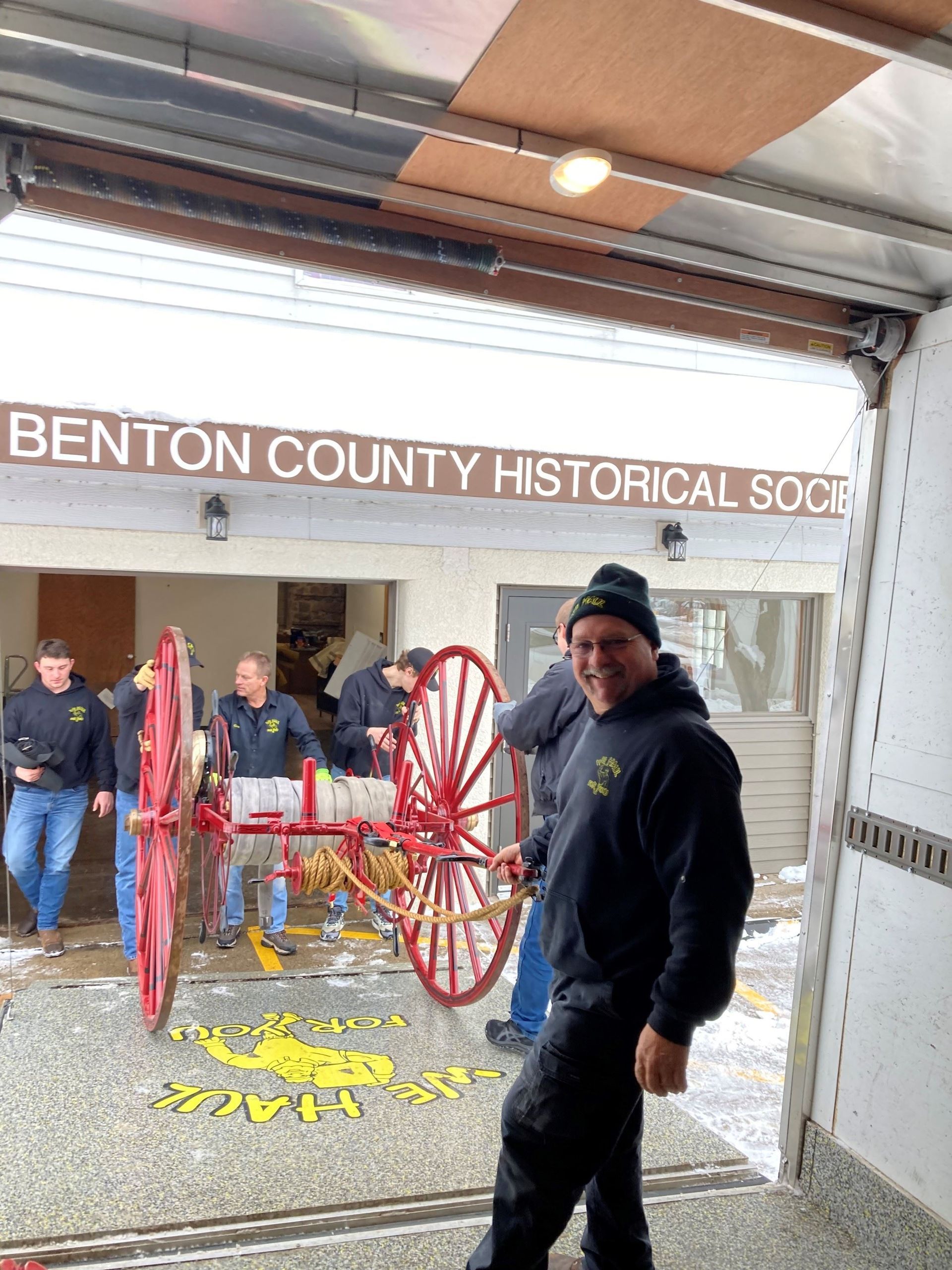 Man smiles in front of a restored red fire wagon. Group in matching jackets at Benton County Historical Society.