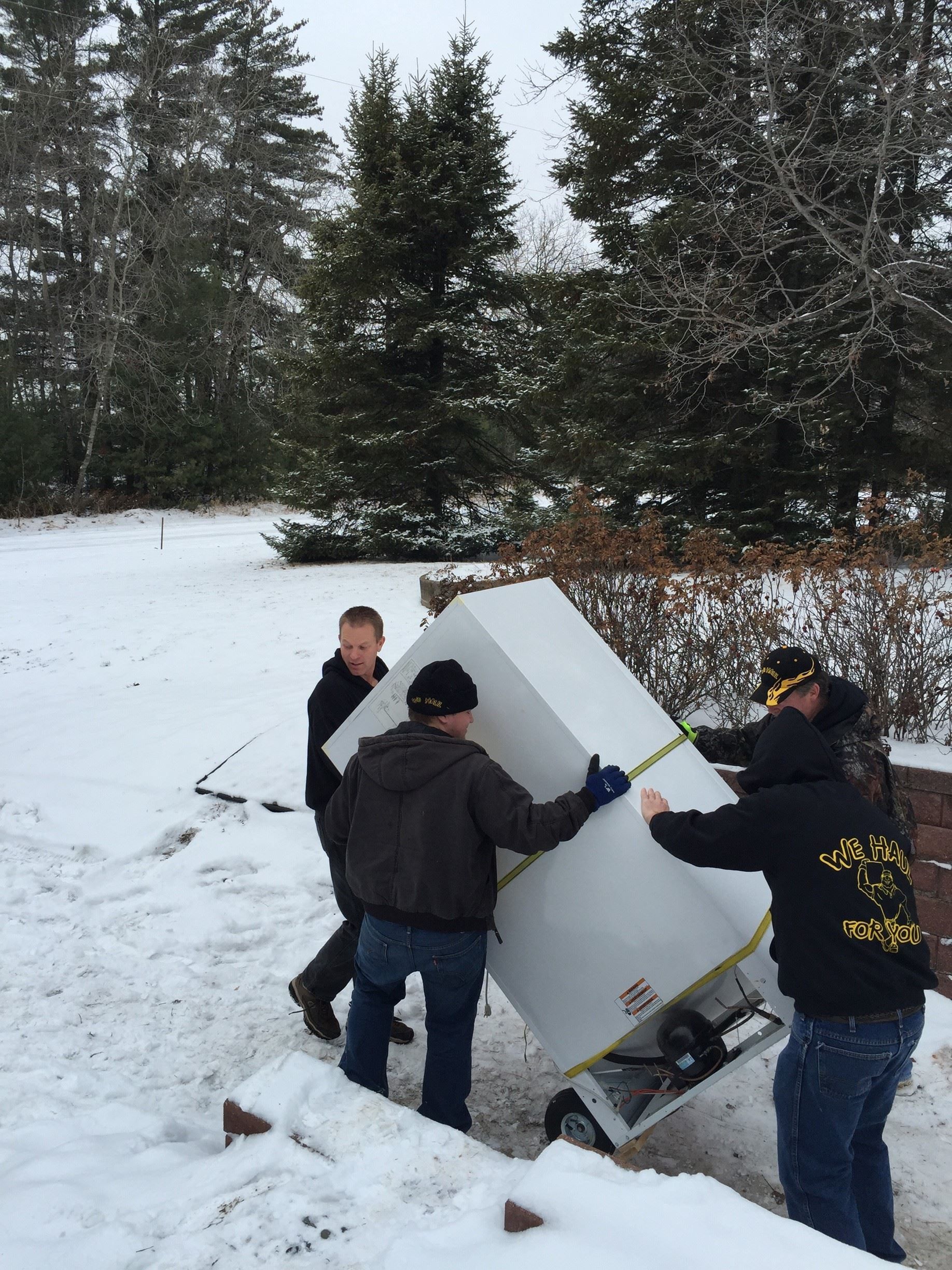 Four people lift a large, white appliance on a dolly in a snowy outdoor setting with trees.