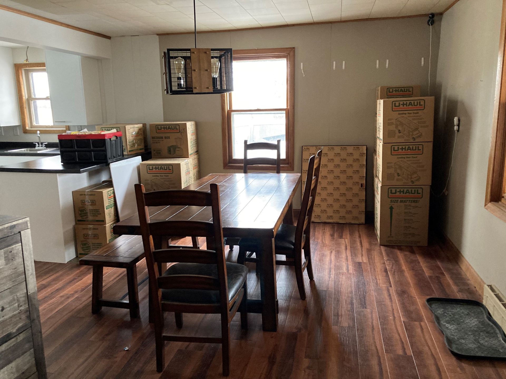 Dining room with a table, chairs, and moving boxes. Dark wood floors. Natural light streams in through a window.