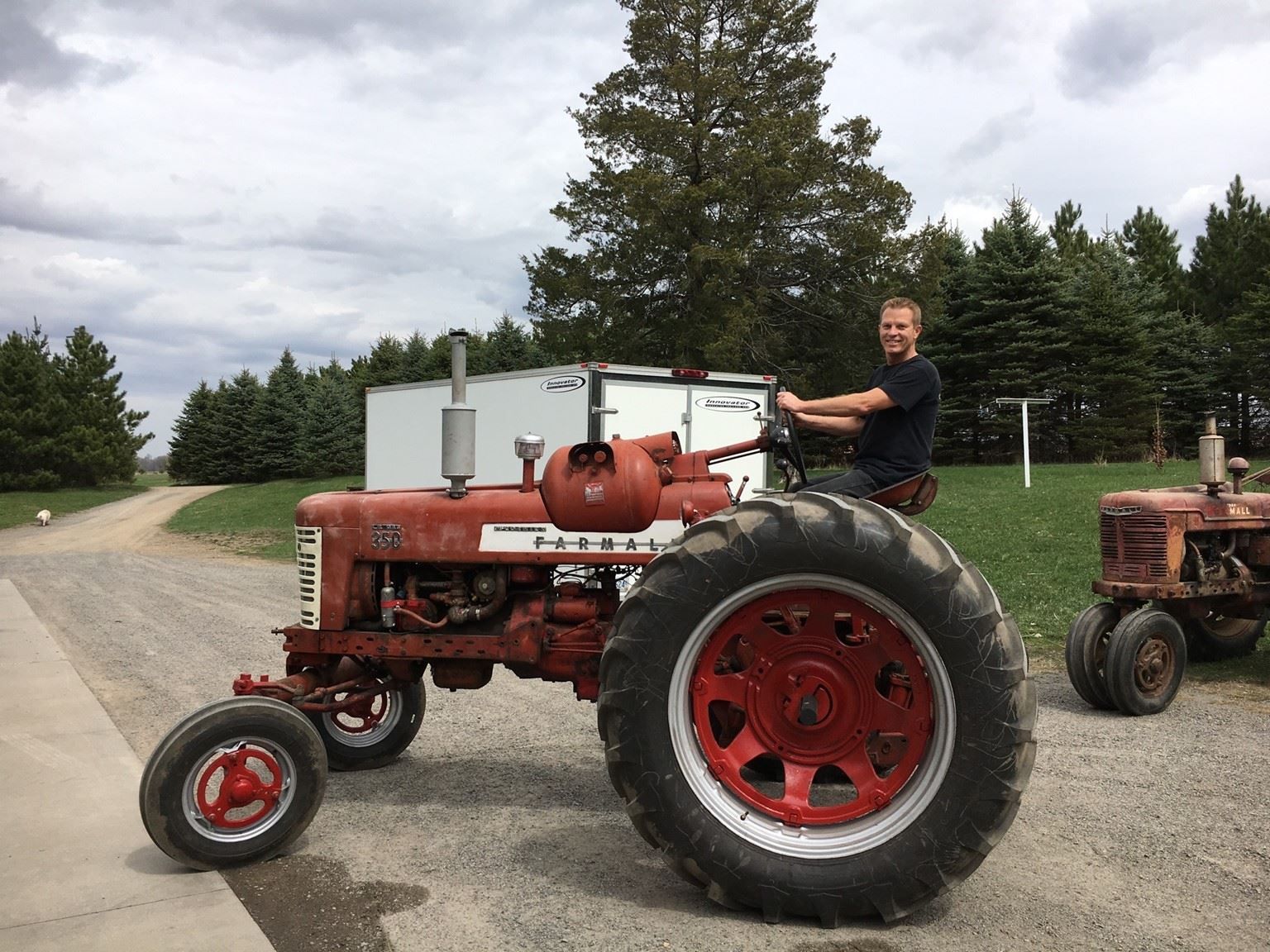Man seated on a red vintage tractor on a farm, with a second tractor nearby.