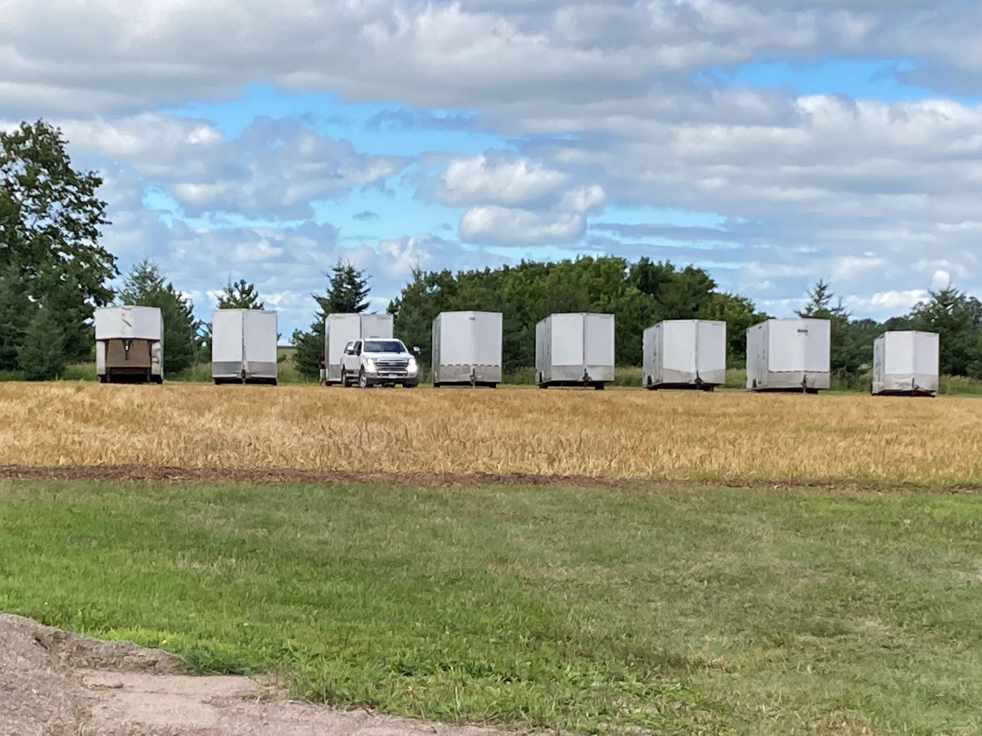 A row of white trailers in a field with a vehicle parked in front of one. Blue sky with clouds.