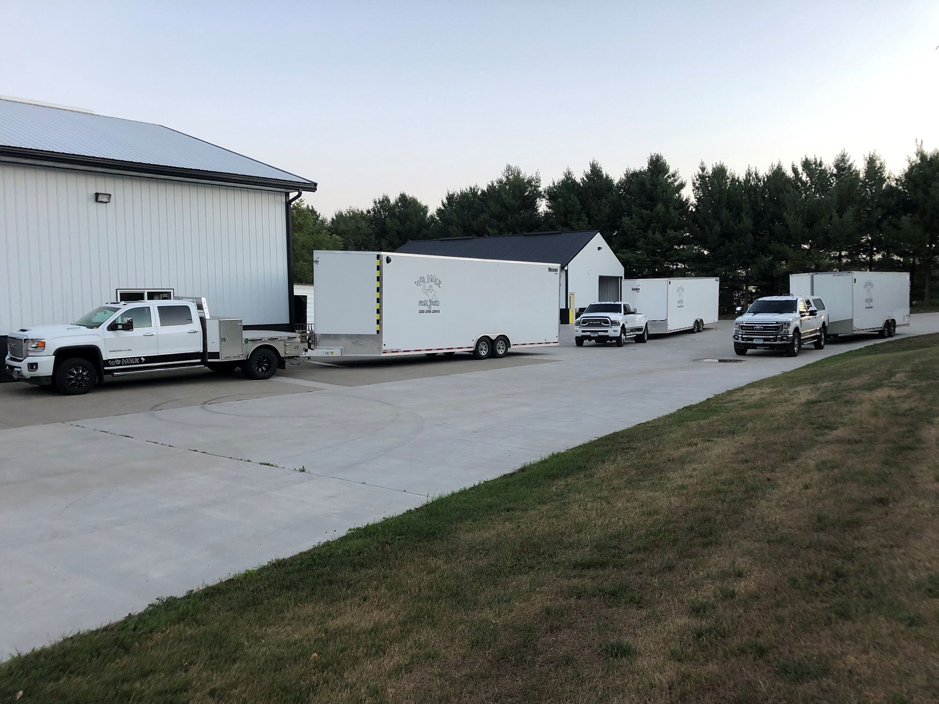 Three white trucks towing trailers parked next to a white building and grass.