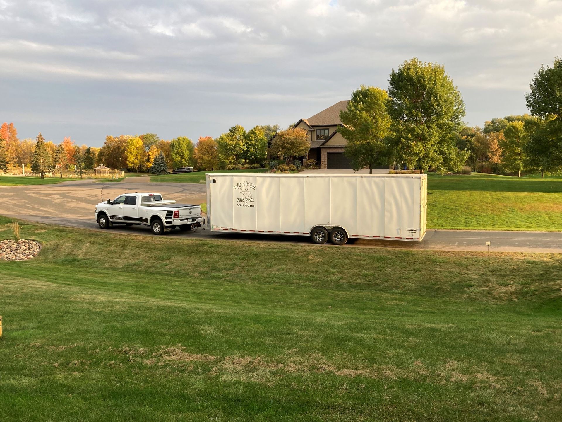 White truck towing a large white trailer on a grassy drive in front of a house, autumn colors.