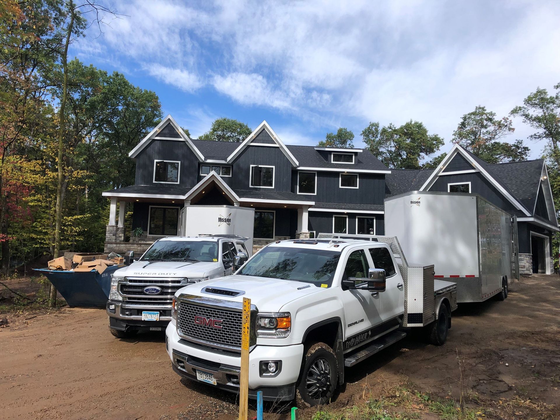 Two white trucks and trailer parked outside a dark gray house under construction.
