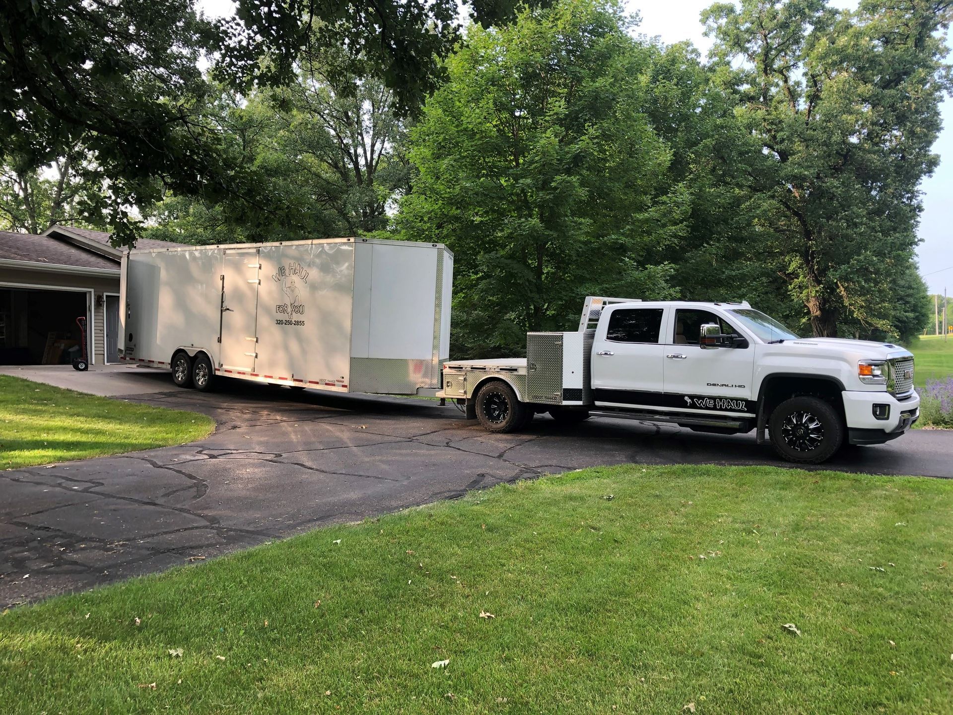 White truck towing a large white trailer on a driveway, green grass, and trees in the background.