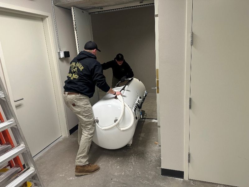 Two people carrying a large white bathtub through a doorway. Interior setting, concrete floors.