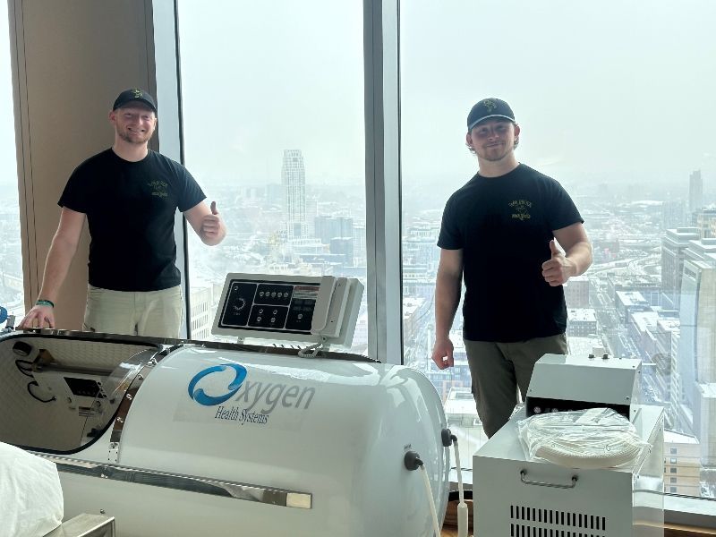 Two men with thumbs up next to a hyperbaric oxygen therapy chamber, overlooking a cityscape.