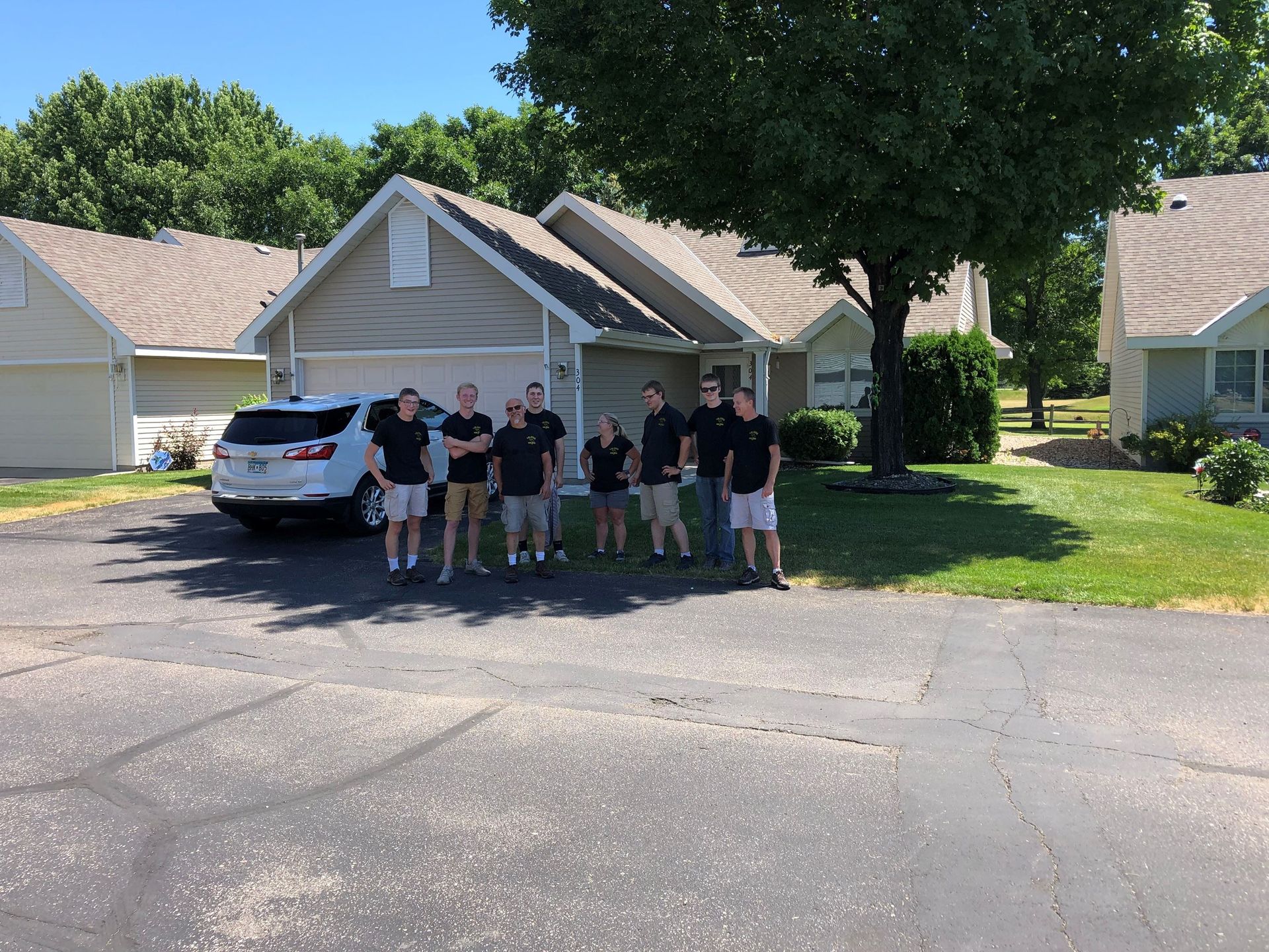 Group of people in black shirts and khaki shorts stand in front of houses with a car parked.