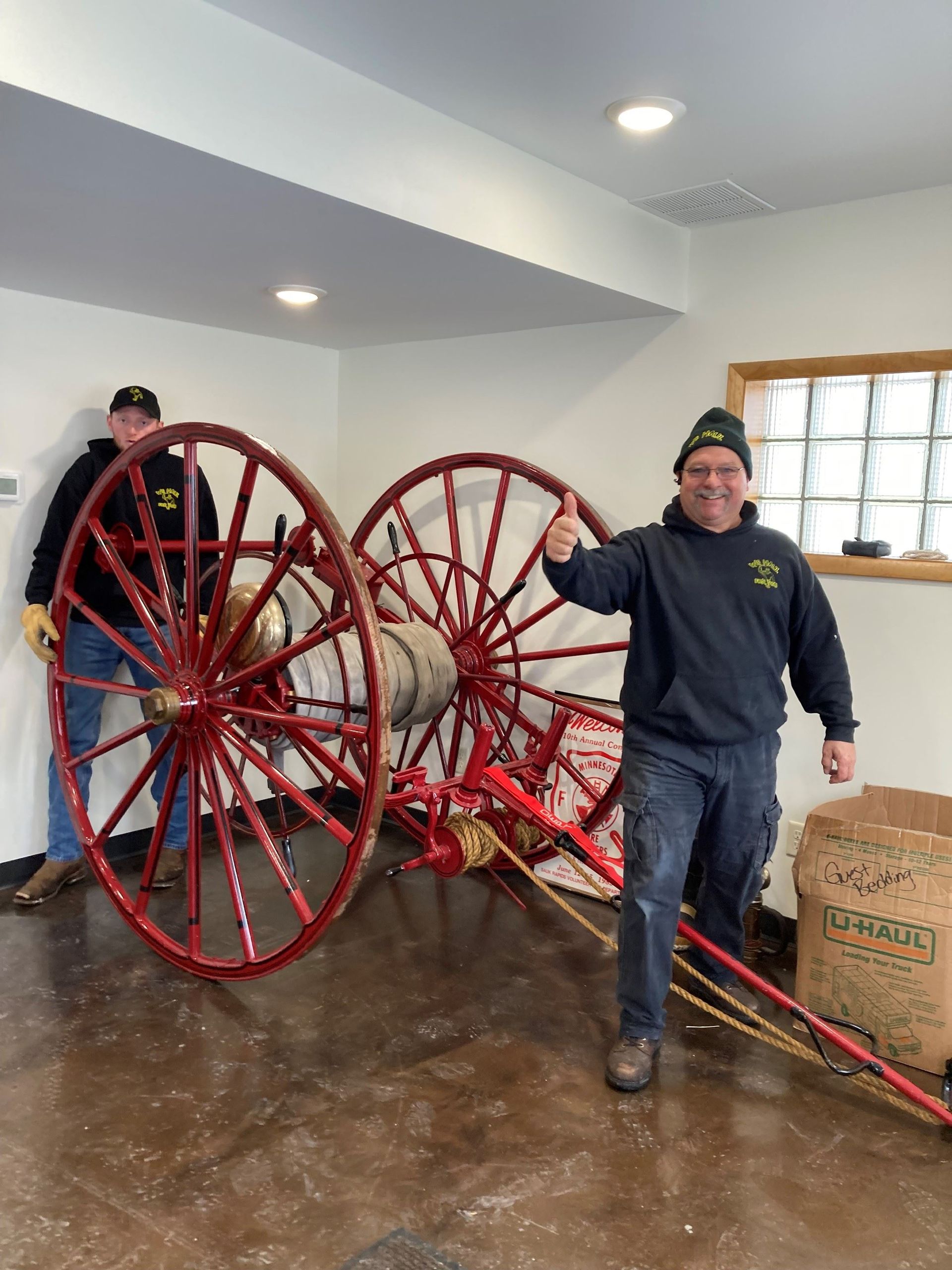 Two men with red vintage fire cart, one giving a thumbs-up. Inside a building.