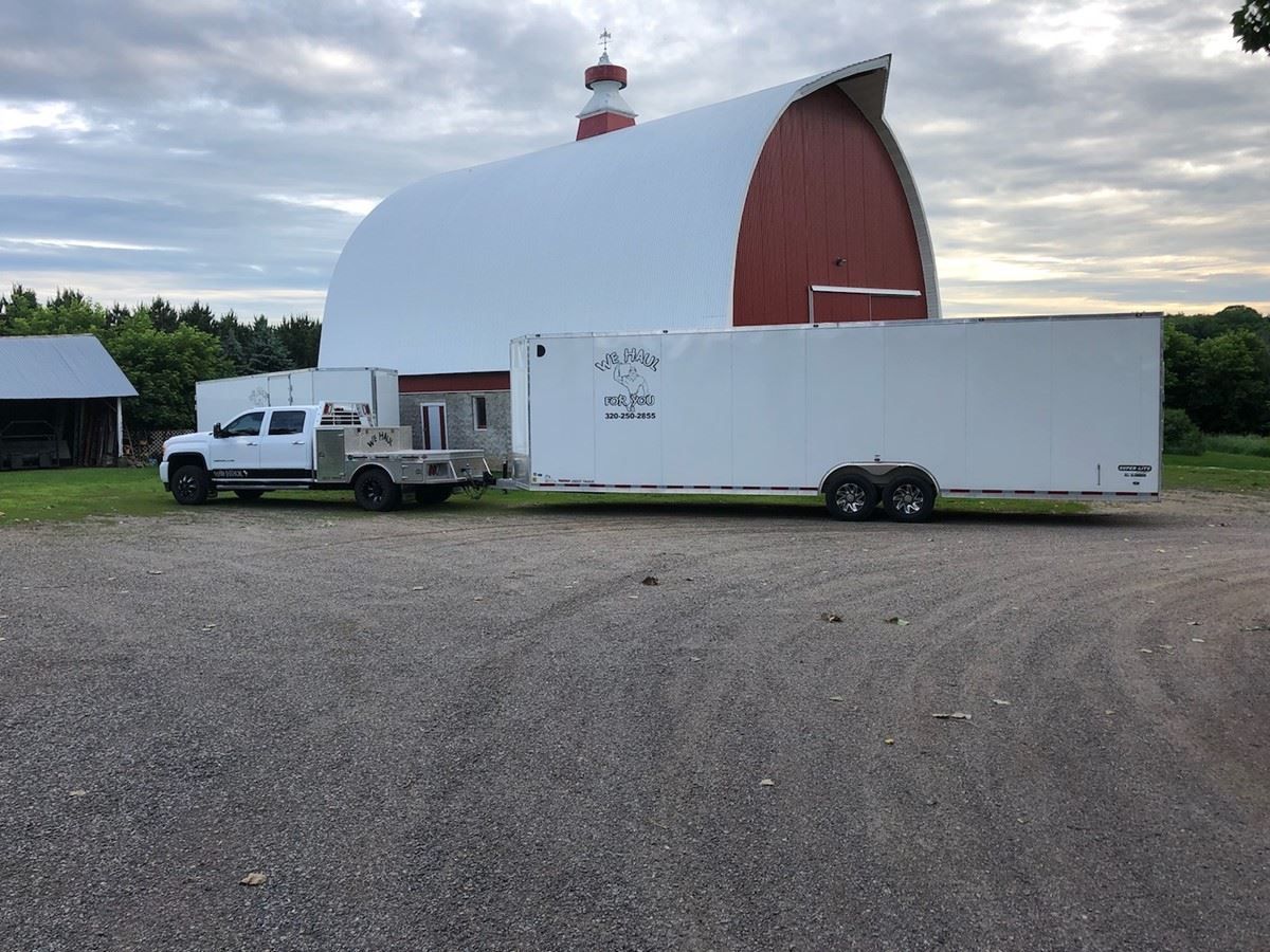 White truck pulling a white trailer parked on gravel in front of a red barn. Overcast sky.