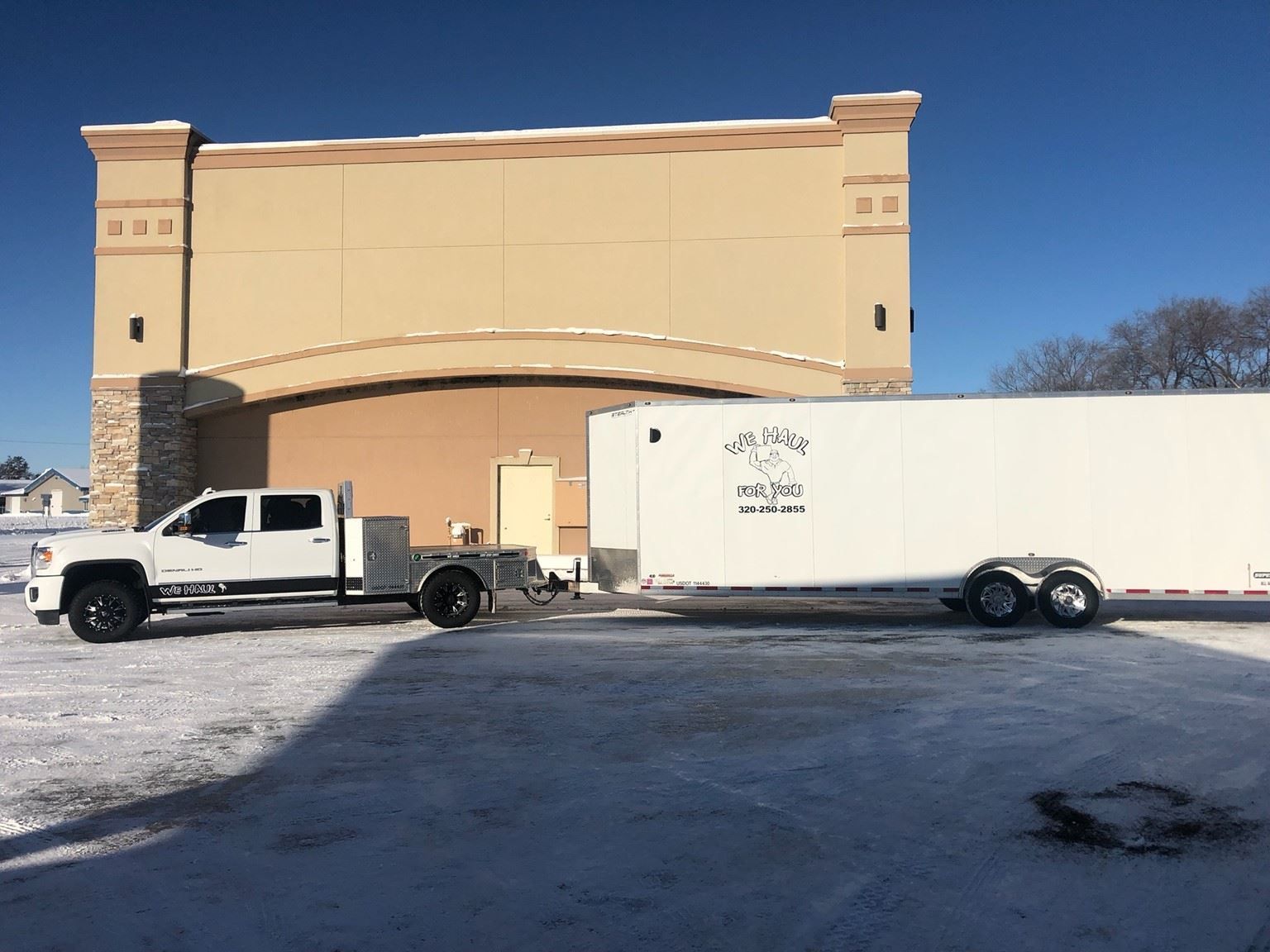 White truck towing a white trailer parked outside a beige building on a snow-covered lot.