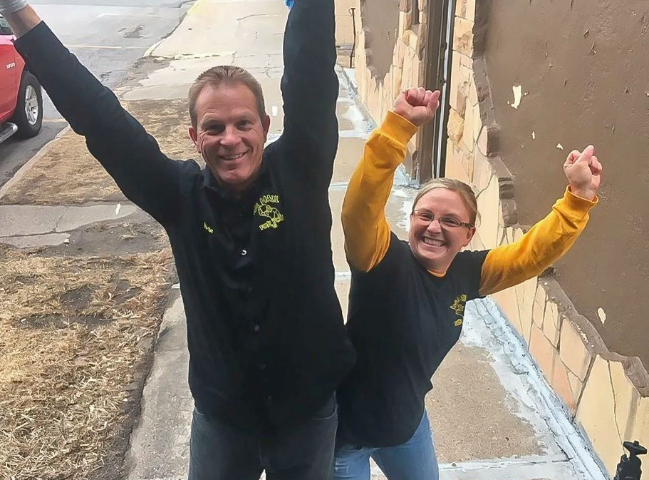Two people with arms raised in front of a building. They wear matching black shirts with a yellow logo, and jeans.