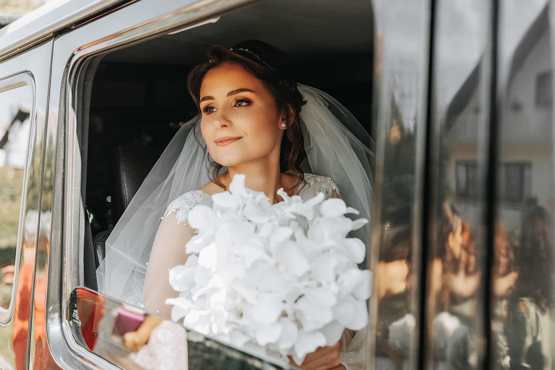 Bride in white dress holding bouquet, looking out car window.