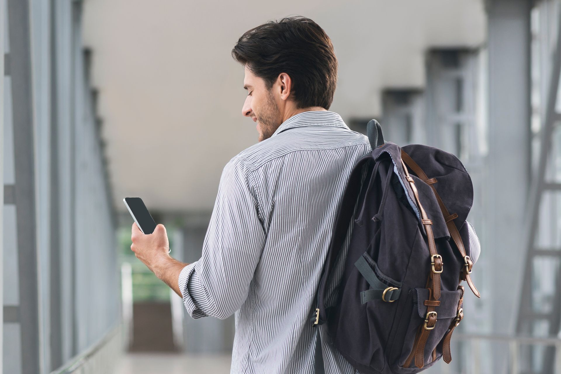 Man with backpack looking at a phone while walking down a hallway.