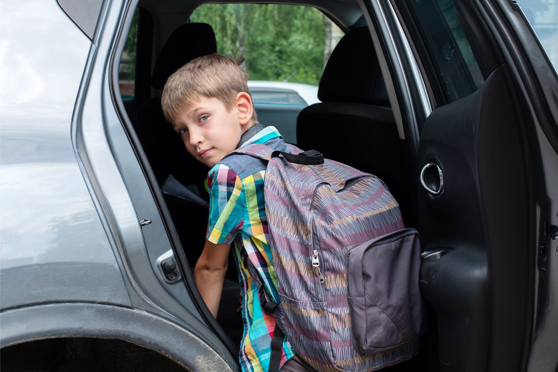 Boy with backpack getting into a gray car. Looking over his shoulder.