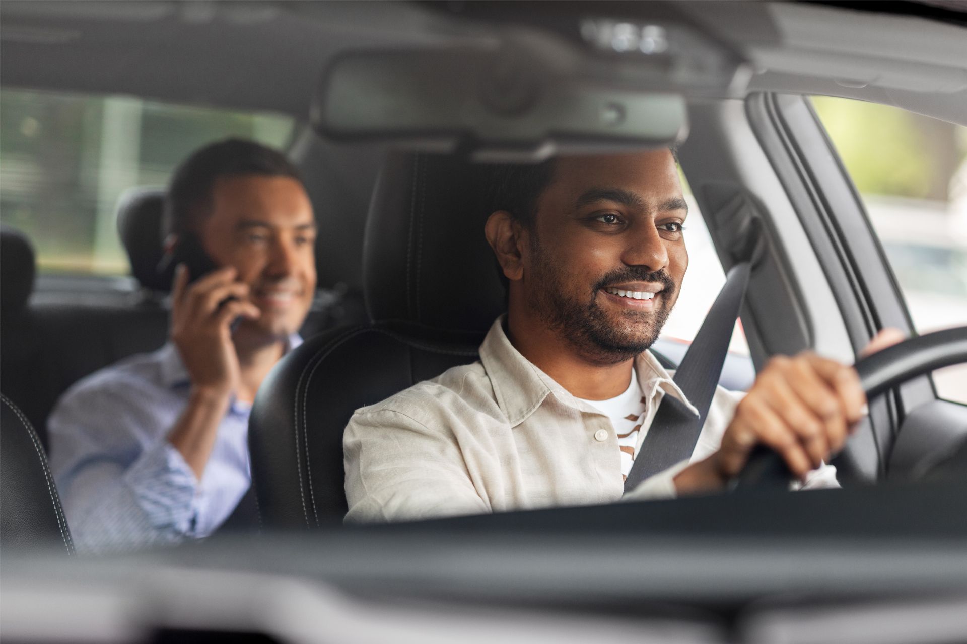 A driver smiles while driving. A passenger in the back speaks on a phone. Inside a car.