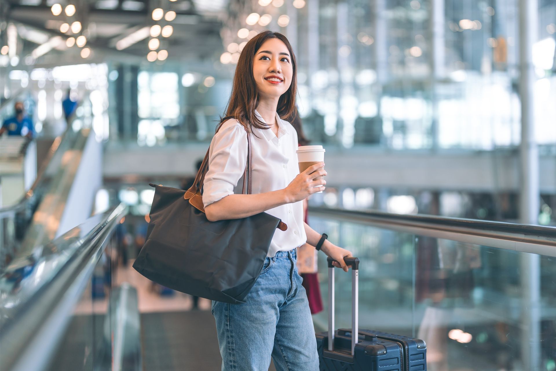 Woman with coffee and luggage in an airport, smiling, looking up, holding a tote bag.