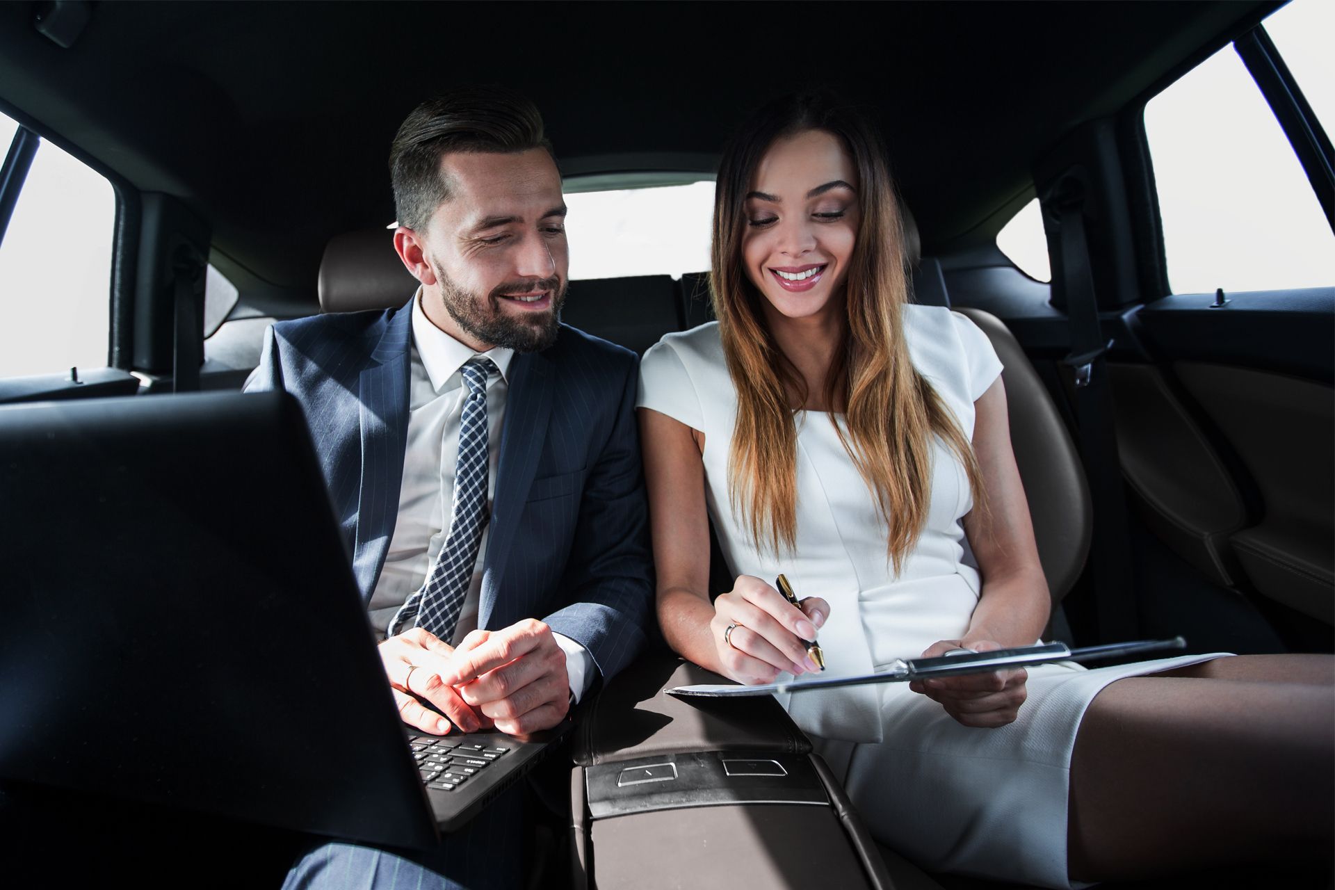 Two professionals working together in the back of a car; man on laptop, woman writing, both smiling.