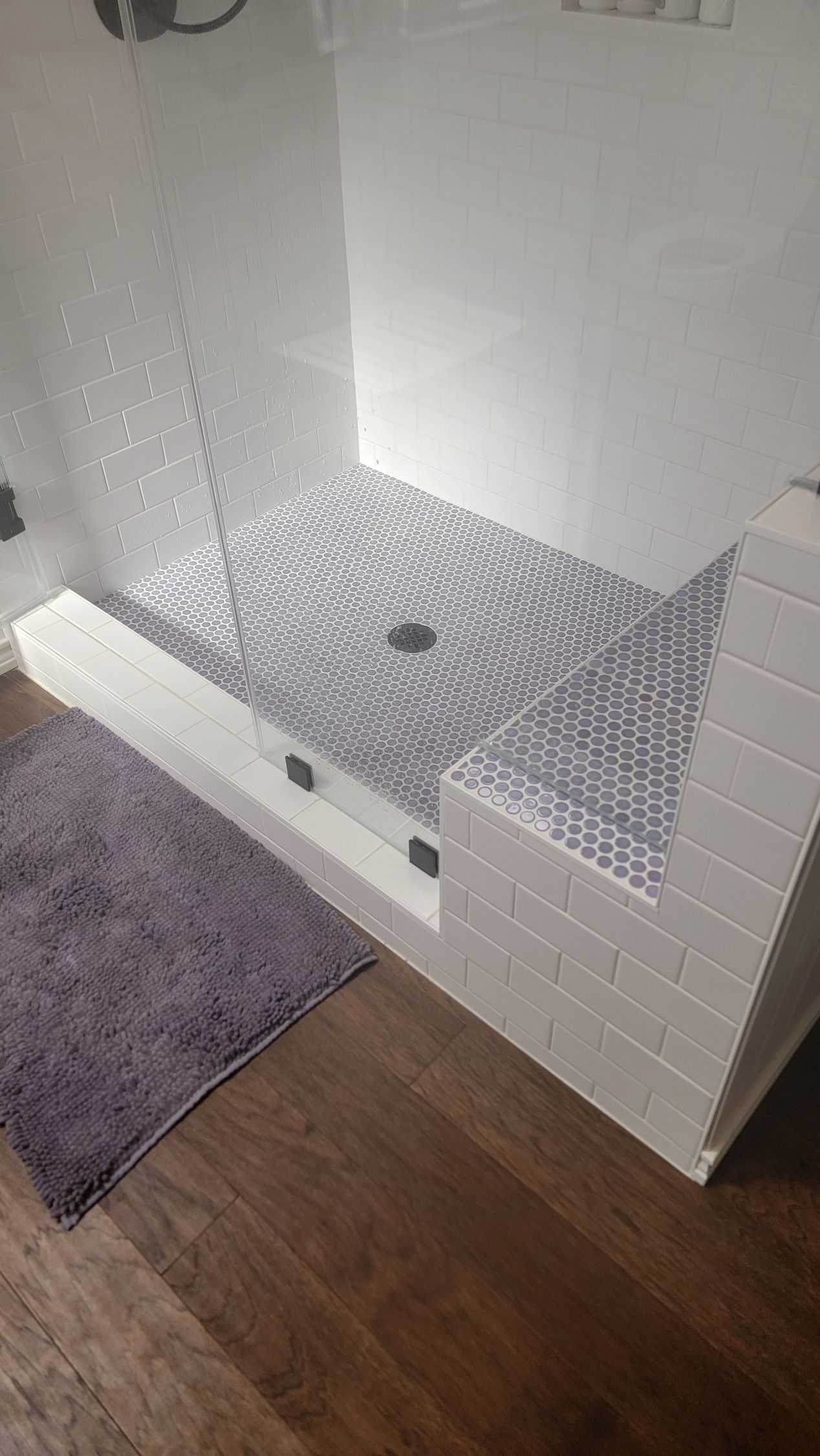 A shower stall with white brick-like walls, a gray and white circular pattern floor, and a purple bath mat.