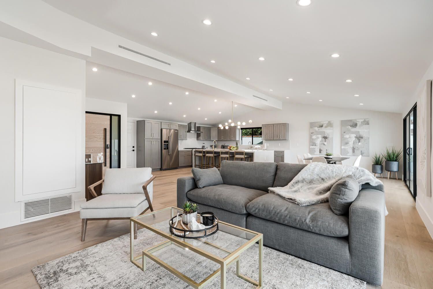 Modern living room with open layout, gray couch, light wood floors, and kitchen in the background.