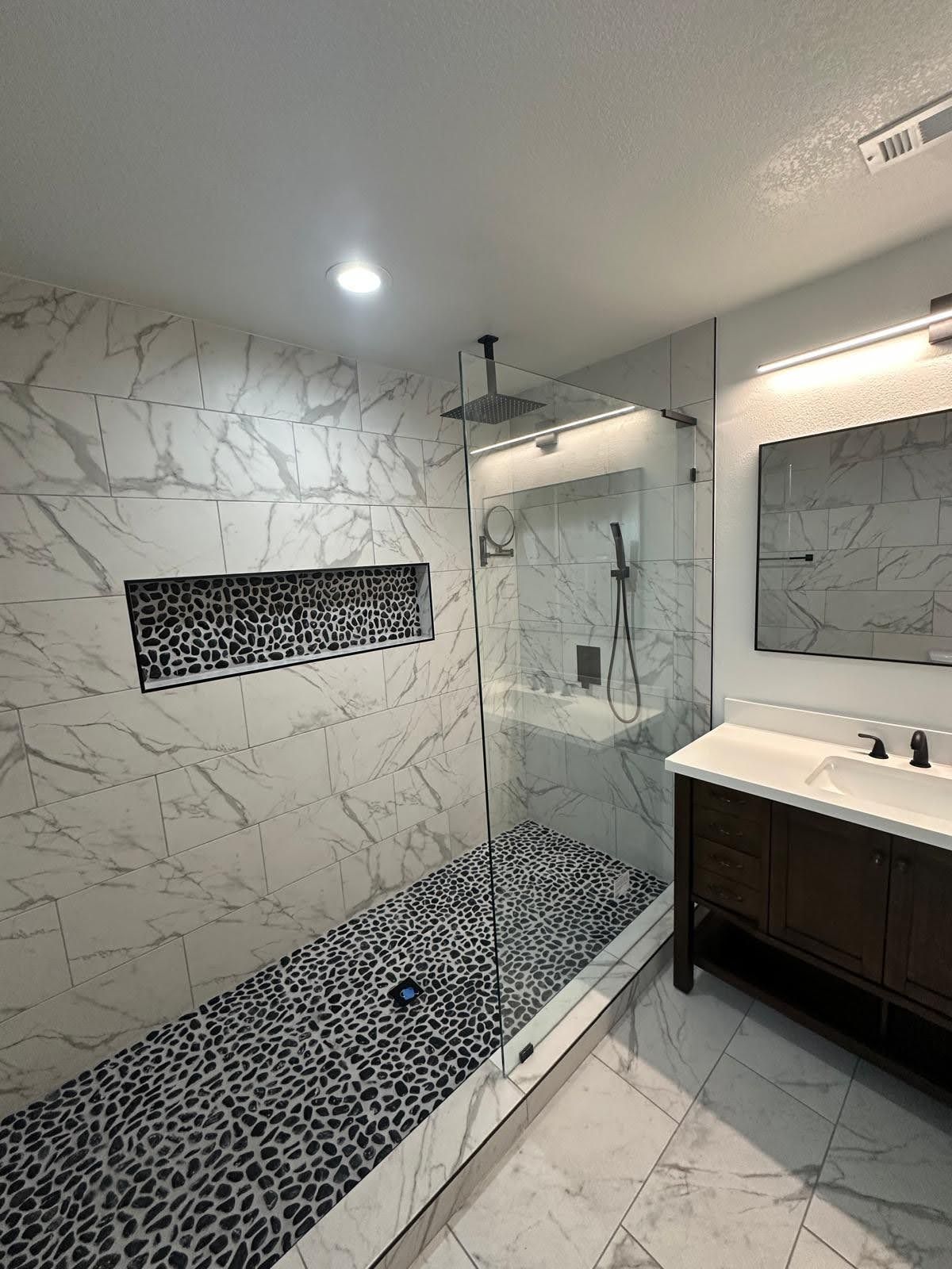 Modern bathroom with marble walls and a pebble floor shower. A dark wood vanity sits under a mirror.
