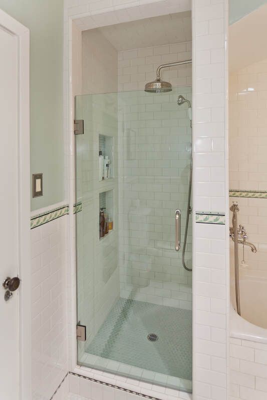 Glass shower enclosure with white tile interior, showerhead, and built-in shelves, next to a bathtub.