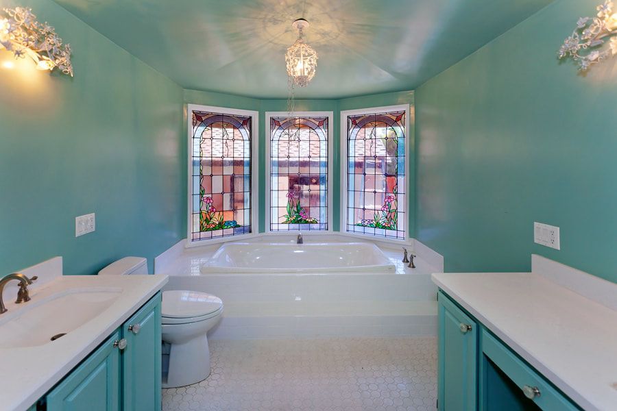 Bathroom with teal walls, white tub, stained-glass windows, and teal cabinets.