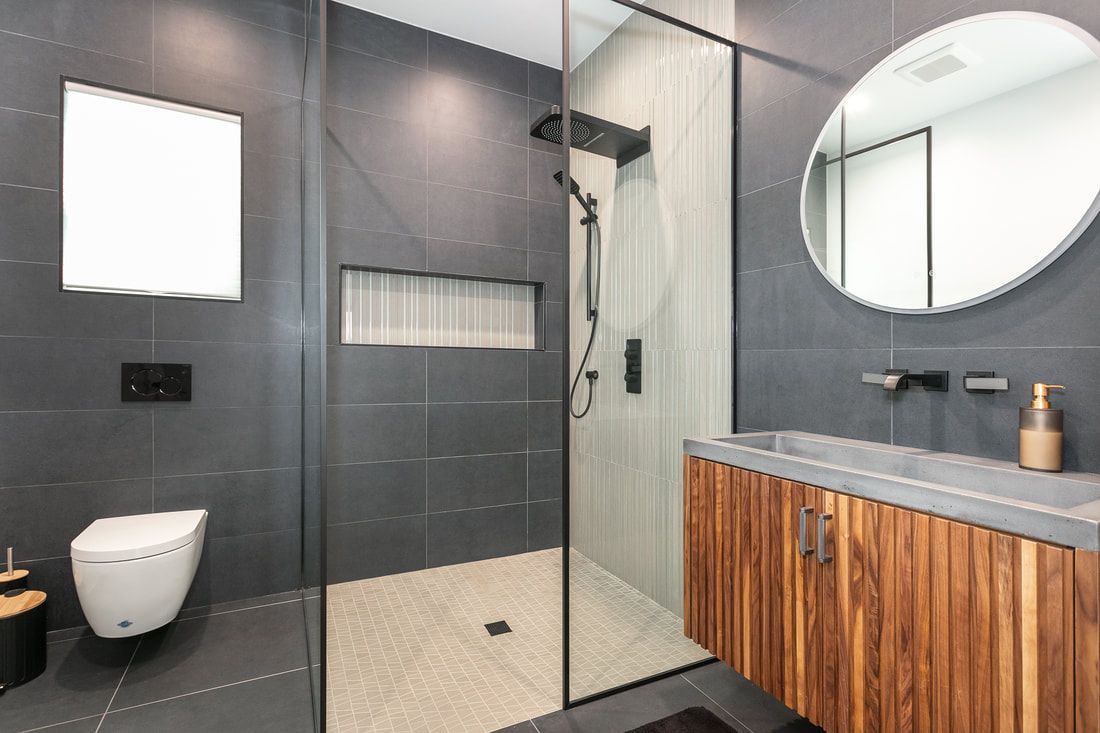 Guest bathroom includes a spacious shower adorned with large dark grey tiles and a wall featuring fluted waterfall glass tiles. It boasts a custom floating walnut vanity with a fluted design, complemented by a concrete sink and a black wall-mounted faucet. The shower is equipped with a three-way black shower head that offers rain, waterfall, and handheld options. The shower enclosure consists of two glass panels with a black frame to complement the black hardware and trim theme.